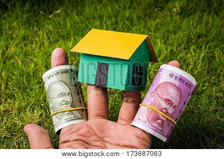 hand holding new indian currency notes or rupees 500 and 2000 and house model over green grass - indian real estate and finance concept