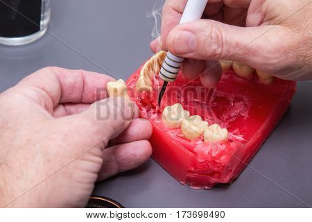 Close up of men's hands working with electric waxer on a red wax dental model on a grey  table