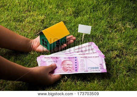 hand holding new indian currency notes or rupees 500 and 2000 and house model over green grass - indian real estate and finance concept