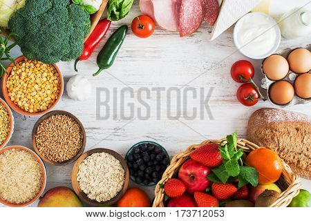 Variety of fruits and vegetables cereals ham cheese eggs bread on the white wooden table top view copy space selecitve focus. Basket of strawberries apples oranges kiwi bowls of oats spelt rice kamut peas; broccoli cauliflower gardlic tomatoes peppers