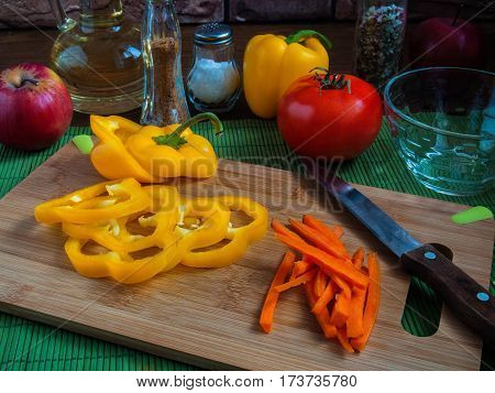Big yellow juicy sliced pepper rings on a wooden cutting board on a kitchen table with carrots red tomatoes apples vegetable oil salt spices