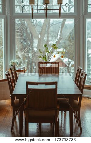 Large classic wooden dining table with six chairs around standing by window lemon tree and flowers on background home interior decoration toned with filters in retro vintage style.