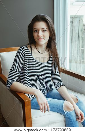 Closeup portrait of smiling Caucasian young beautiful woman model with messy long hair in ripped blue jeans and striped t-shirt sitting armchair by window indoor looking in camera toned with filters