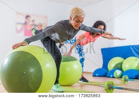 Fit Caucasian women doing crane exercise standing on one leg with their arms out to sides holding on to balance ball while training in fitness club.