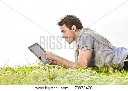 Side view of young man using digital tablet while lying on grass against clear sky