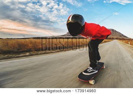 A young man in a fullface helmet and a leather suit in a special rack rides a longboard on afsaltu sunset in the background mountains and beautiful sky at high speed.