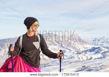 Young beautiful girl in sunglasses with Nordic poles in the sunset light on the background of the Caucasian ridge and rock