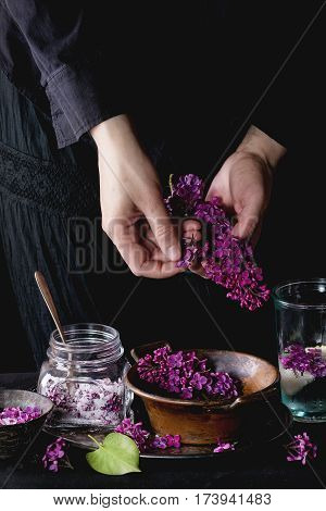 Female hands  pluck lilac flowers from bunch to vintage copper bowl. Glass jar of lilac flowers in sugar and glass of lilac water with lemon on black tablecloth over black. Dark rustic atmosphere.