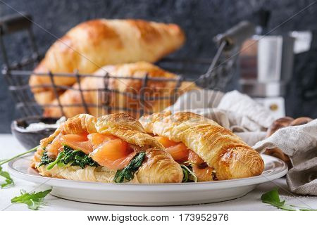 Croissant with smoked salted salmon, spinach and arugula served on white plate with bowl of cream cheese and coffee maker over white and gray concrete background. Close up