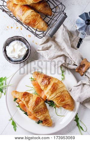 Croissant with smoked salted salmon, spinach and arugula served on white plate with bowl of cream cheese and coffee maker over white texture concrete background.