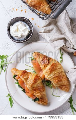 Croissant with smoked salted salmon, spinach and arugula served on white plate with bowl of cream cheese and coffee maker over white texture concrete background.