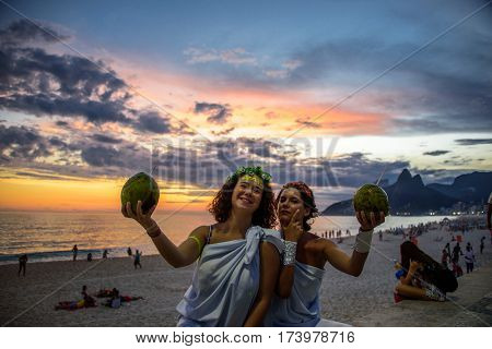 RIO DE JANEIRO, BRAZIL - FEBRUARY 27, 2017: Two women in the costumes of Greek Goddesses wearing flowers diadems and holding coconuts on the background of the beautiful sundown at Ipanema beach