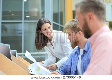 Group Of Young People In Casual Wear Sitting At The Office Desk And Discussing Something While Looki