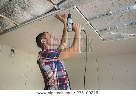 Young Man In Usual Clothing And Work Gloves Fixing Drywall Suspended Ceiling To Metal Frame Using El