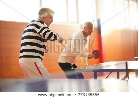 Two active senior men with rackets attacking ping pong ball while standing by tennis table during game