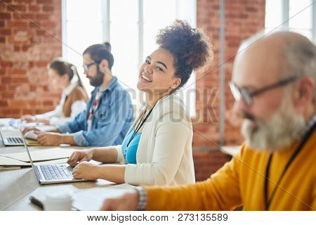 Young cheerful mixed-race businesswoman in casualwear looking at you with smile among her co-workers