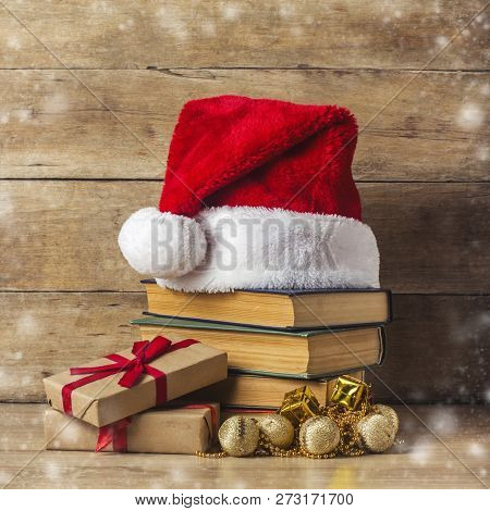 A Stack Of Books, Santa Clauss Hat, Gift Boxes On A Wooden Background. Concept Of New Year And Merry