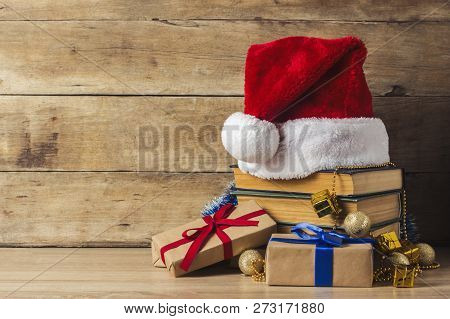 A Stack Of Books, Santa Clauss Cap, Gift Boxes With Red And Blue Ribbons On A Wooden Background. Con