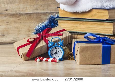 A Stack Of Books, Santa Clauss Cap, An Alarm Clock, Gift Boxes On A Wooden Background. Concept Of Ne