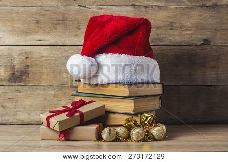 A Stack Of Books, Santa Clauss Cap, Christmas-tree Decorations, Gift Boxes On A Wooden Background. C