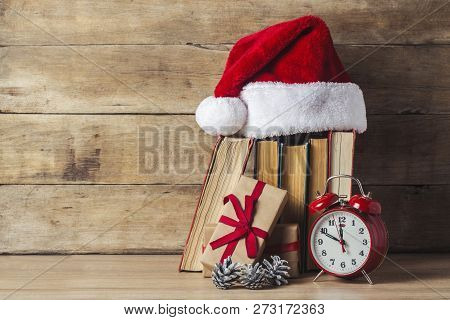 A Stack Of Books, Santa Clauss Cap, An Alarm Clock, Cones, Gift Boxes On A Wooden Background. Concep