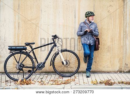 Active Senior Man With Electrobike Standing Outdoors In Town, Using Smartphone.