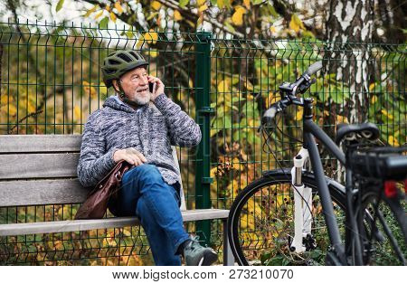 Senior Man With Electrobike Sitting On A Bench Outdoors In Town, Using Smartphone.