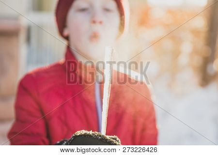 Young Boy In Red Jacket, Hoodie, And Gloves Holding An Icicle And Blowing Breath On It.