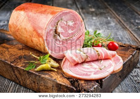 Traditional Allgauer veal roast roll sausage sliced and as piece with tomatoes and chili as closeup on an old rustic cutting board