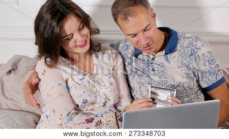Charming Pregnant Woman With Man Sitting With Paper Fetal Scant And Laptop Excited With Childbirth
