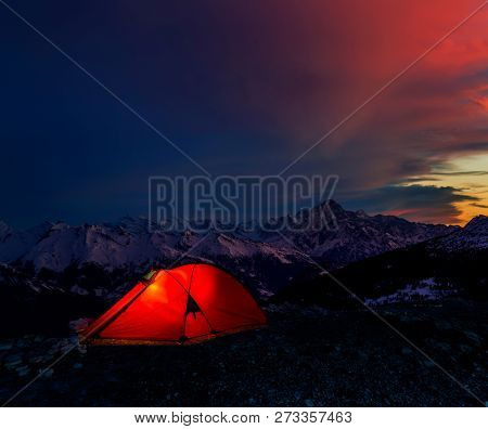 Night bivouac in Mountains, milion star hotel under night sky, red illuminated tent on pass in Alps.