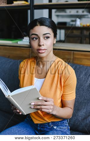 Beautiful Mixed Race Girl In Orange Shirt Holding Book In Living Room And Looking Away