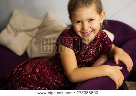 Portrait Of Little Happy Smiling Girl In Shiny Bordo Colour Dress On Bordo Clouor Sofa