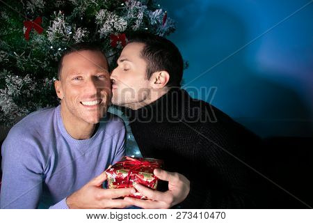 Gay Male Couple Exchanging Christmas Gift In Front Of Tree With A Kiss
