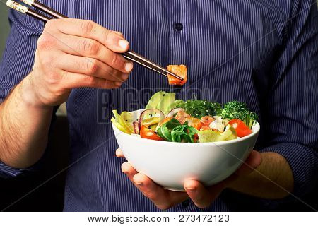 Man In A Shirt Holds Poke Bowl With Salmon, Avocado, Cucumbers, Arugula, Broccoli, Rice, Carrots, An