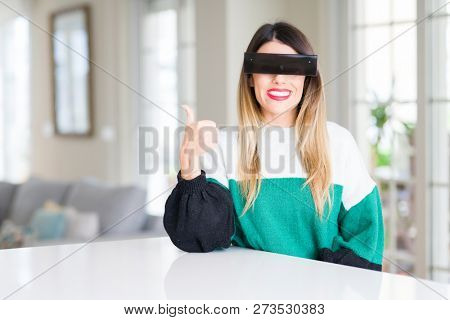 Young beautiful woman blindfold eyes with black censor band at home doing happy thumbs up gesture with hand. Approving expression looking at the camera with showing success.