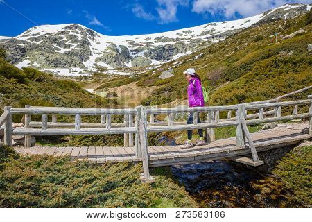 Woman Mountaineer Crossing A Bridge Over A Stream Looking At Mountain