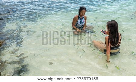 Mother And Daughter With Mobile Phone Sitting In Sea At Beach Surrounded By Tropical Fish