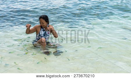 Asian Woman Sitting In Water At Beach, Laughging And Playing While Surrounded By Tropical Fish