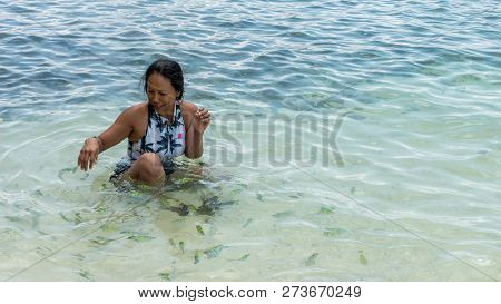 Asian Woman Sitting In Water At Beach, Laughging And Playing While Surrounded By Tropical Fish