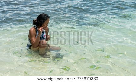 Asian Woman Sitting In Water At Beach, Laughging And Playing While Surrounded By Tropical Fish