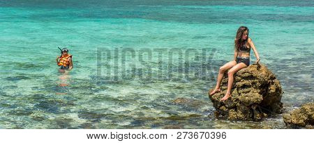 Mother In Water With Mask And Snorkle While Daughter Sits On Rock In Two Piece Bathing Suit