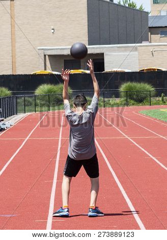 A High School Track Runner Is Throwing A Medicine Ball Over His Head Backwards On A Red Track.