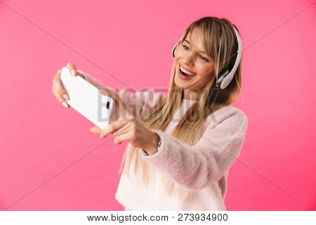Cheerful young blonde woman wearing sweater standing isolated over pink background, listening to music with headphones, taking a selfie