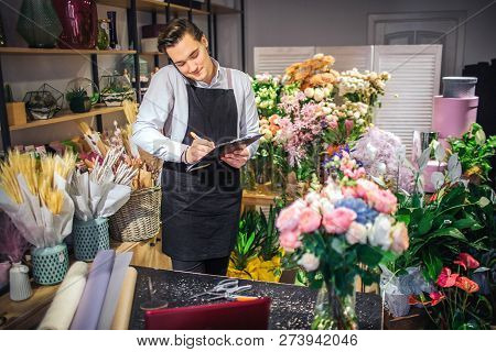 Picture Of Young Male Florist Talk On Phone And White Down On Black Planchette. He Has Laptop, Vase 