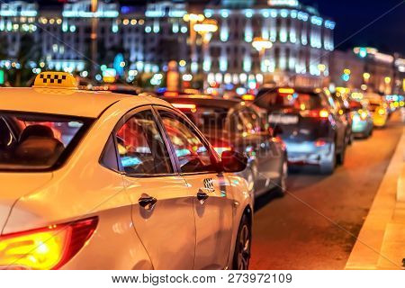 Taxi In Traffic Jam Among Street Cars In The Evening Rush Hour In Megalopolis On Background Of Light