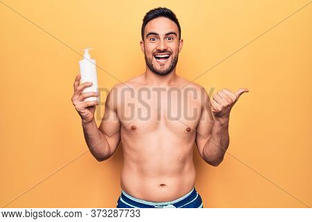 Handsome man with beard on vacation wearing swimwear holding bottle of sunscreen lotion pointing thumb up to the side smiling happy with open mouth