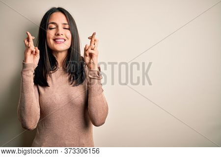 Young brunette woman with blue eyes wearing casual sweater over isolated white background gesturing finger crossed smiling with hope and eyes closed. Luck and superstitious concept.