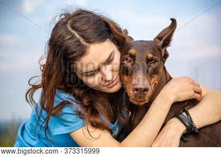 Girl Lovingly Hugs Her Doberman Dobermann Dog And Looks At Him. Portrait Against The Sky. Horizontal