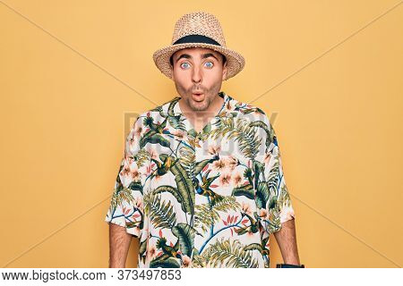 Young handsome man with blue eyes on vacation wearing summer florar shirt and hat afraid and shocked with surprise expression, fear and excited face.